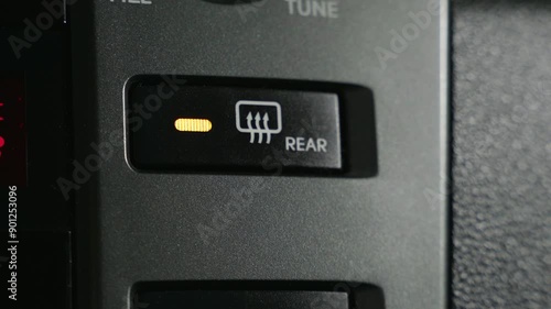 A close-up view of a rear window defroster button inside a car. The button is black and has a white icon of a window with three arrows pointing upwards. The word REAR is printed below the icon