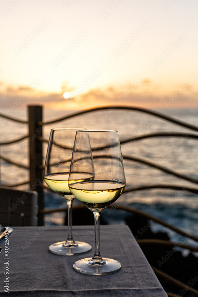 Obraz premium Romantic outdoor evening with two glasses of white wine and beautiful sunset on the sea beach at background. Italy, Sicily, evening time, summer. Vertical image.