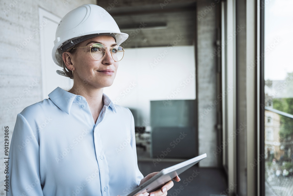 Thoughtful engineer wearing hardhat standing with tablet PC in office Thoughtful engineer wearing hardhat standing with tablet PC in office