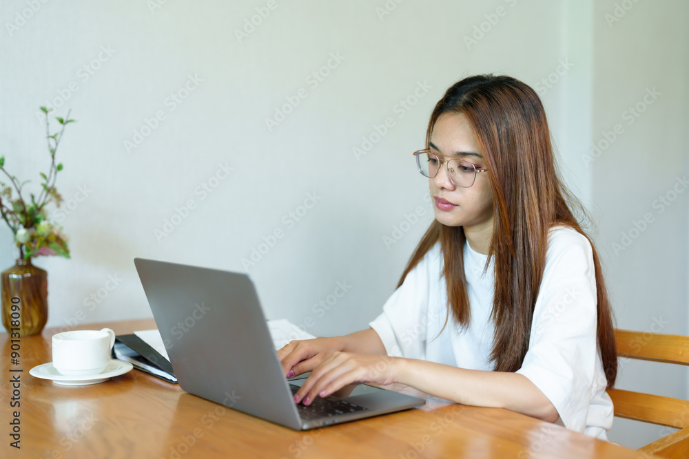 Asian woman wearing glasses sitting at wooden table working on laptop, white cup and saucer beside, casual attire, indoor home office setting, daytime. Bright natural light Asian woman wearing glasses sitting at wooden table working on laptop, white cup and saucer beside, casual attire, indoor home office setting, daytime. Bright natural light