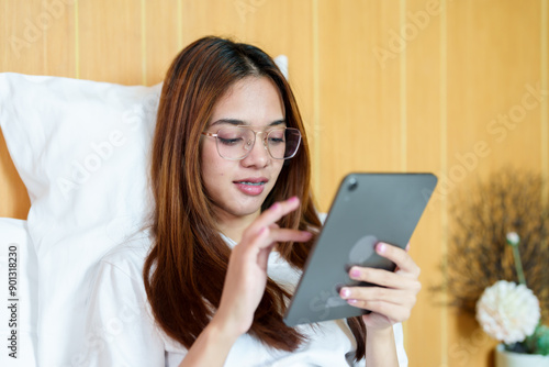 Beautiful Asian woman with glasses sitting in bed, holding a tablet, watching a movie. Wearing casual white t-shirt, relaxed posture, Indoor bedroom setting, comfortable and cozy atmosphere.