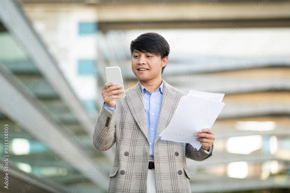 Asian businessman in a gray plaid blazer stands in a modern city setting, holding papers in one hand and using his smartphone with the other. The background is an urban environment. Asian businessman in a gray plaid blazer stands in a modern city setting, holding papers in one hand and using his smartphone with the other. The background is an urban environment.