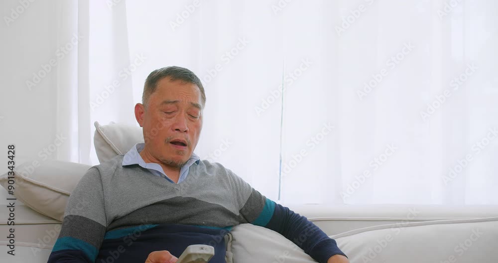 Senior Elderly Asian man sitting comfortably on Couch holding a TV remote Control in Bright Living Room with White Curtains Senior Elderly Asian man sitting comfortably on Couch holding a TV remote Control in Bright Living Room with White Curtains
