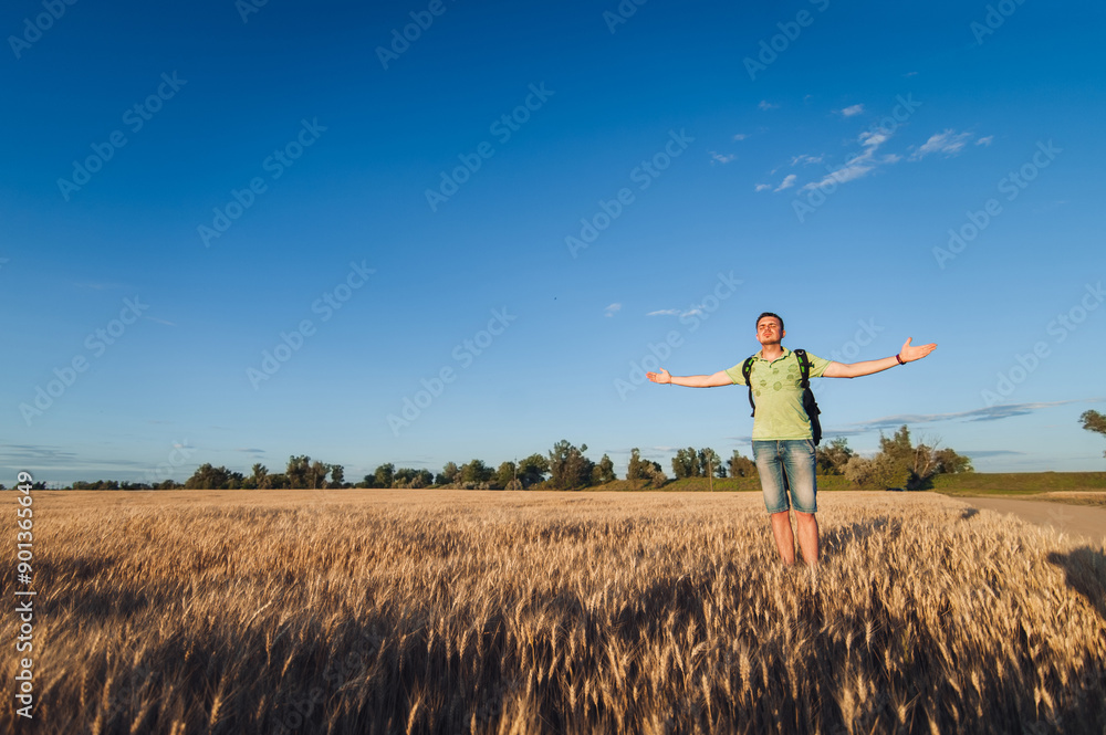 A young man in a T-shirt, shorts and a backpack stands on a hill in a yellow wheat field and spreads his arms wide, feeling free. Peace and idyll. Countryside. The setting sun shines on the man's face A young man in a T-shirt, shorts and a backpack stands on a hill in a yellow wheat field and spreads his arms wide, feeling free. Peace and idyll. Countryside. The setting sun shines on the man's face