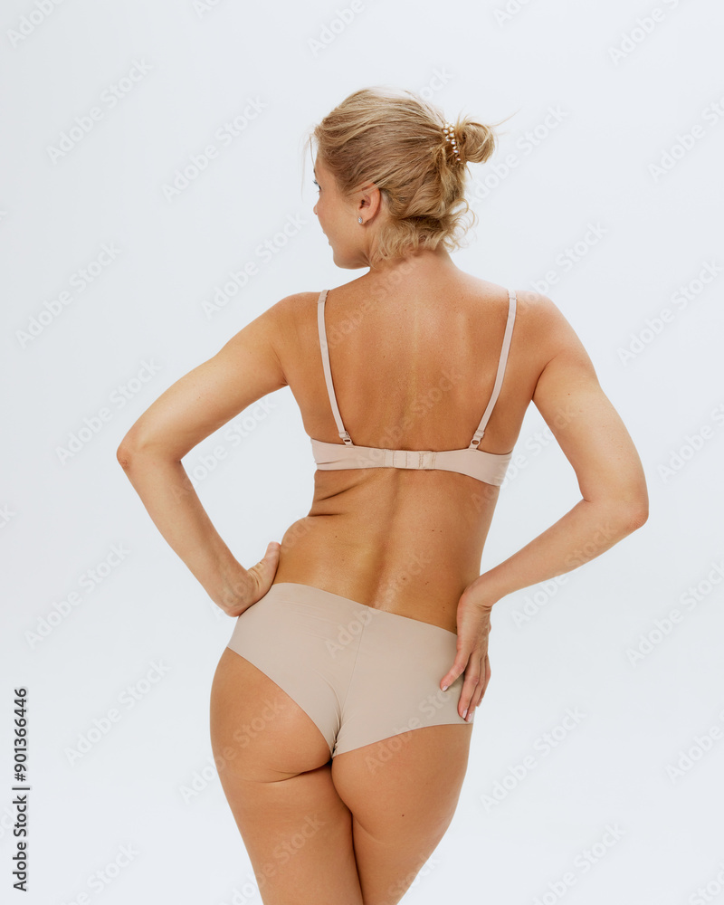 Naklejka premium Rear view of blonde woman in beige lingerie posing holding hands on hips against white studio background. Concept of beauty and female health, lifting, cosmetology procedures, body care, antiaging. Ad