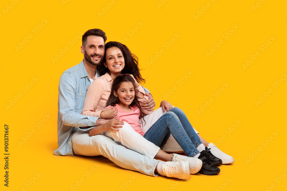 A family of three, a father, a mother, and their daughter, are sitting together on a bright yellow background. They are all smiling and looking at the camera. The father has his arm around the mother A family of three, a father, a mother, and their daughter, are sitting together on a bright yellow background. They are all smiling and looking at the camera. The father has his arm around the mother
