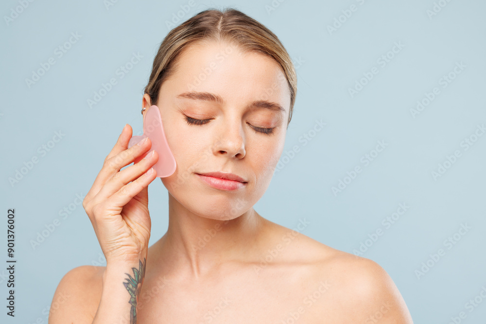 Half naked topless calm young woman with nude make up doing face massage holding rose quartz guache scraper isolated on plain blue color background. Skin care healthcare cosmetic procedures concept. Half naked topless calm young woman with nude make up doing face massage holding rose quartz guache scraper isolated on plain blue color background. Skin care healthcare cosmetic procedures concept.