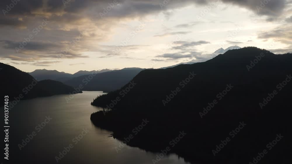 Silhouette of evergreen forest and mountain peaks with orange glow on clouds, aerial descend