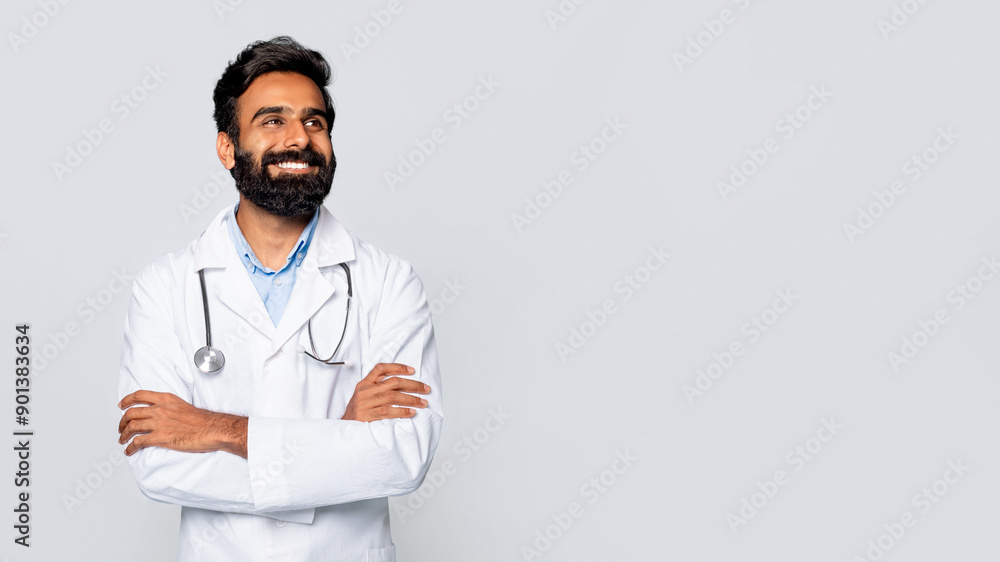 A smiling Indian man doctor wearing a white lab coat and stethoscope stands in front of a plain white background, arms crossed, looking at copy space A smiling Indian man doctor wearing a white lab coat and stethoscope stands in front of a plain white background, arms crossed, looking at copy space