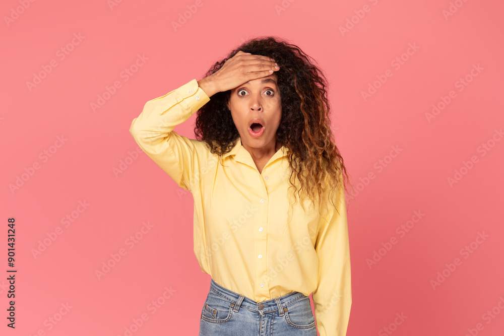 A young black woman with curly brown hair is wearing a light yellow button-up shirt and blue jeans. She is standing in front of a pink background and looks surprised with her mouth open wide A young black woman with curly brown hair is wearing a light yellow button-up shirt and blue jeans. She is standing in front of a pink background and looks surprised with her mouth open wide