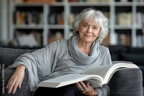 Elderly woman with gray hair reading a large open book while sitting on a gray couch in a library with shelves of books in the background