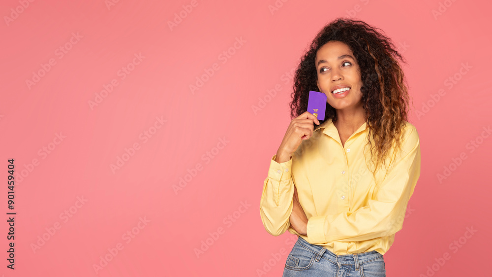 African American young woman with curly hair is wearing a yellow shirt and blue jeans. She is smiling and holding a purple card to her chin as she looks at copy space
