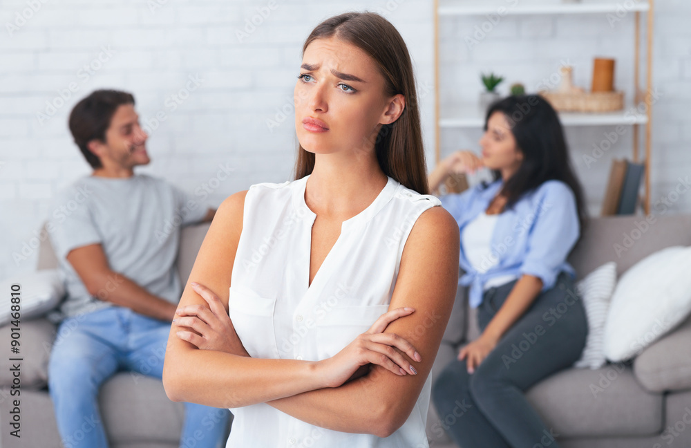 Jealousy. Sad Girl Standing Offended While Her Boyfriend Flirting With Girl Sitting On Couch Indoor. Selective Focus Jealousy. Sad Girl Standing Offended While Her Boyfriend Flirting With Girl Sitting On Couch Indoor. Selective Focus