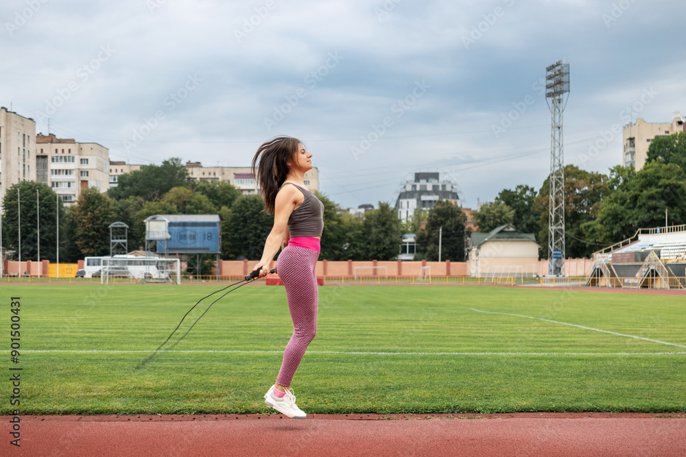 Obraz premium A young sporty woman doing her morning workout, skipping rope on the stadium, outdoors, side view