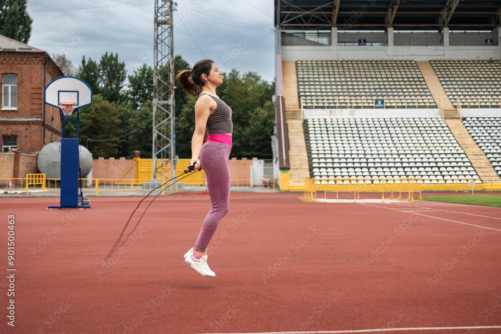Obraz premium A young sporty woman doing her morning workout, skipping rope on the stadium, outdoors, side view