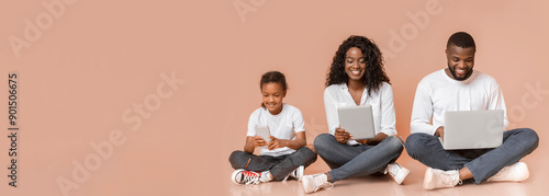 Modern family. Happy african american father, mother and daughter using different gadgets while sitting on floor over yellow studio background, panorama