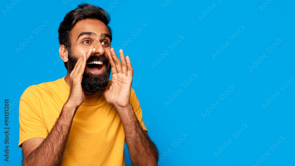 Indian young man with a beard is standing in front of a blue background. He is cupping his hand around his mouth and shouting, copy space. He has a yellow t-shirt on and appears happy and excited. Indian young man with a beard is standing in front of a blue background. He is cupping his hand around his mouth and shouting, copy space. He has a yellow t-shirt on and appears happy and excited.