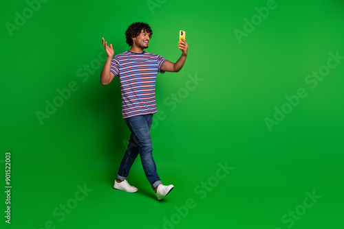 Portrait of young indian curly funny man student in striped t shirt waving hand making creative video isolated on green color background © deagreez