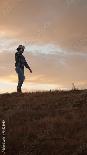 A Cowboy, Silhouetted, is Walking on a Grassy Hill at Sunset under the Western Skyline
