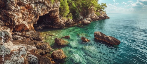 High-quality photo of a rocky shore lined with green trees, featuring a cave in the rock, and clear blue water in the seascape with abundant copy space image.