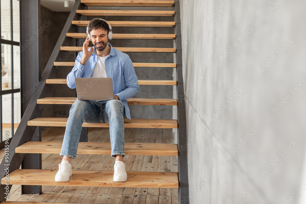 Fototapeta premium A man sits on a wooden staircase with headphones on, working on a laptop while smiling. He is wearing blue jeans and a light blue button-down shirt, copy space