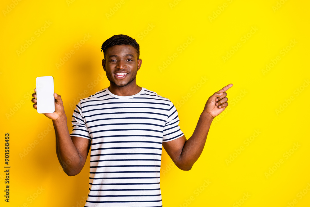Photo of nice young man point finger show empty space phone screen wear striped t-shirt isolated on bright yellow color background Photo of nice young man point finger show empty space phone screen wear striped t-shirt isolated on bright yellow color background