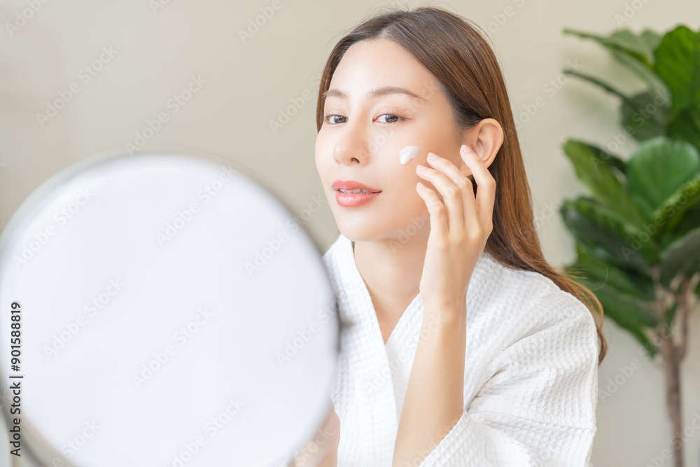 Facial beauty skin care, smile of pretty asian young woman in bathrobe looking at mirror, hand applying moisturizer lotion on her face, holding jar of skin cream before makeup cosmetic routine at home Facial beauty skin care, smile of pretty asian young woman in bathrobe looking at mirror, hand applying moisturizer lotion on her face, holding jar of skin cream before makeup cosmetic routine at home