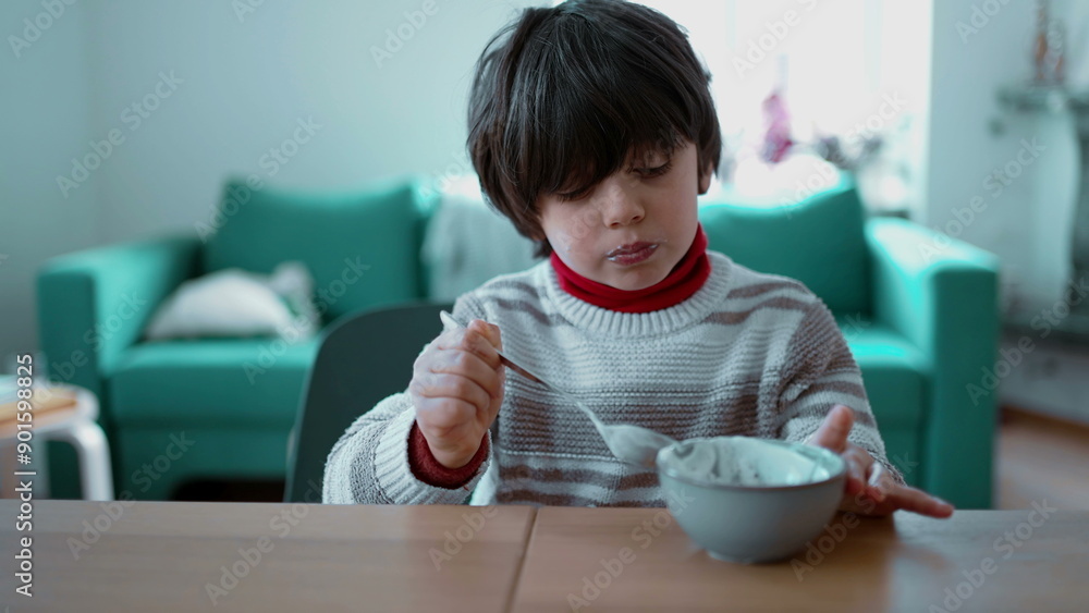 Young boy eats yogurt with a spoon while seated at the table, focusing on each spoonful, capturing a moment of quiet enjoyment in a cozy home setting with a comfortable sofa Young boy eats yogurt with a spoon while seated at the table, focusing on each spoonful, capturing a moment of quiet enjoyment in a cozy home setting with a comfortable sofa