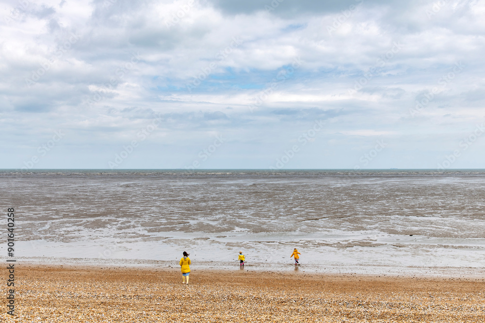 Obraz premium Woman and two children in bright yellow rain gear on pebblestone beach and coastline with mudflats and horizon near Littlestone-on-Sea, Kent, UK with white clouded sky