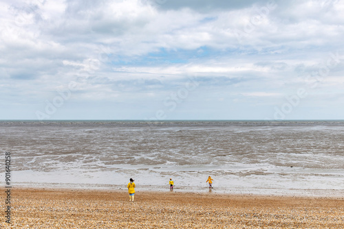 Woman and two children in bright yellow rain gear on pebblestone beach and coastline with mudflats and horizon near Littlestone-on-Sea, Kent, UK with white clouded sky photo