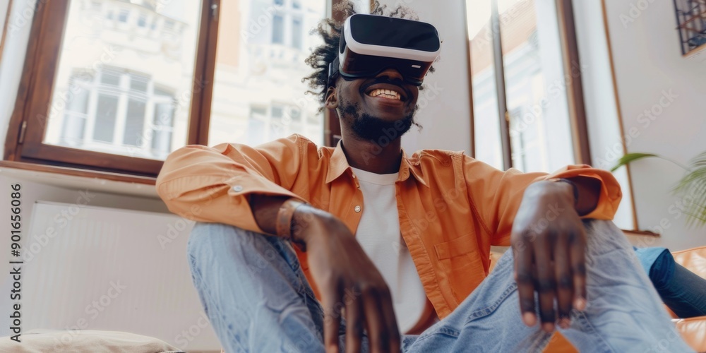 Fototapeta premium A man in an orange shirt is sitting on a couch with his hands on his knees. He is wearing a virtual reality headset and he is smiling