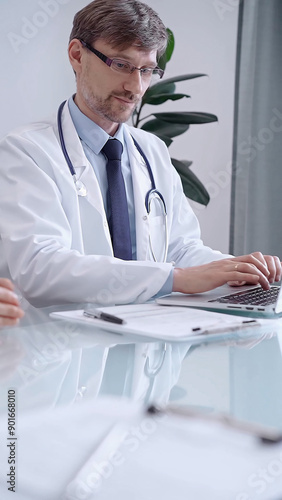 Male physician in white coat focused on laptop data in a modern clinic office while analyzing patient information on laptop. Medicine concept