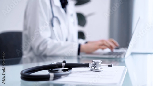 Stethoscope is lying on the glass table while doctor woman is working with laptop computer on the background. Medicine and pharmacy concept