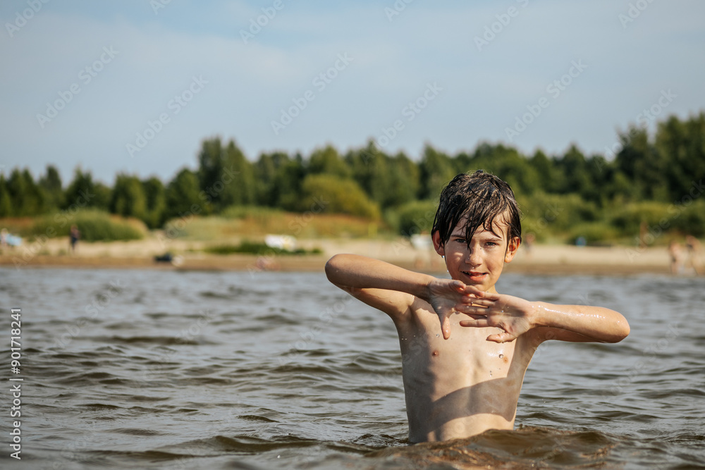 Fototapeta premium A smiling boy with arms outstretched stands in the water at a beach. Other beachgoers are visible in the background under a clear sky