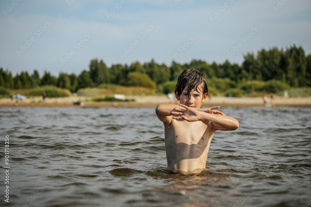 Naklejka premium A smiling boy with arms outstretched stands in the water at a beach. Other beachgoers are visible in the background under a clear sky