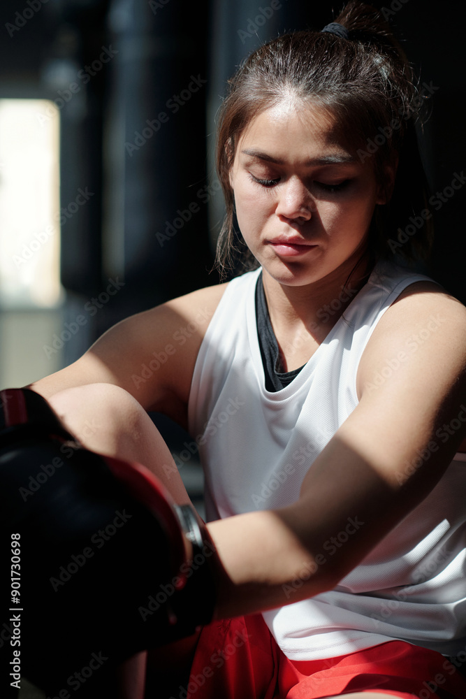 Restful girl in white vest and leather boxing gloves enjoying break between trainings while sitting in front of camera in sunlit gym Restful girl in white vest and leather boxing gloves enjoying break between trainings while sitting in front of camera in sunlit gym