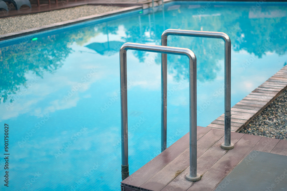 Obraz premium Metal pool ladder leading into a clear blue swimming pool with wooden decking and gravel surround