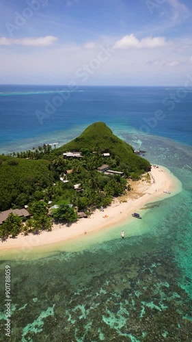 Aerial view over remote Fijian village and resort on small pacific island covered in white sand, palm trees and shallow turquoise water. 