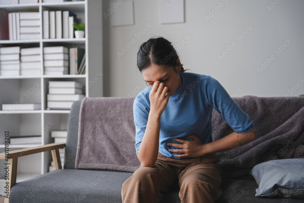 Young woman is sitting on a sofa at home, experiencing discomfort and holding her stomach with a pained expression Young woman is sitting on a sofa at home, experiencing discomfort and holding her stomach with a pained expression