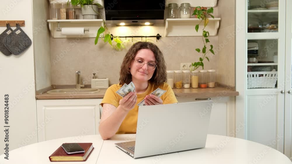 A woman with a laptop holds money dollars at a table in a home kitchen. An adult female businesswoman works from home, a remote office A woman with a laptop holds money dollars at a table in a home kitchen. An adult female businesswoman works from home, a remote office