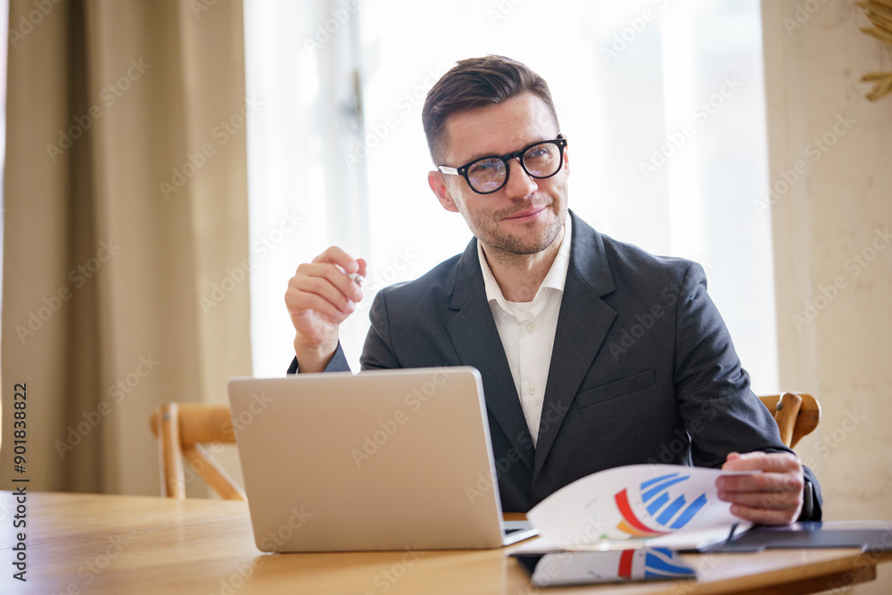 A businessman in glasses and a suit reviews charts at a desk with a laptop, smiling and thoughtful. A businessman in glasses and a suit reviews charts at a desk with a laptop, smiling and thoughtful.