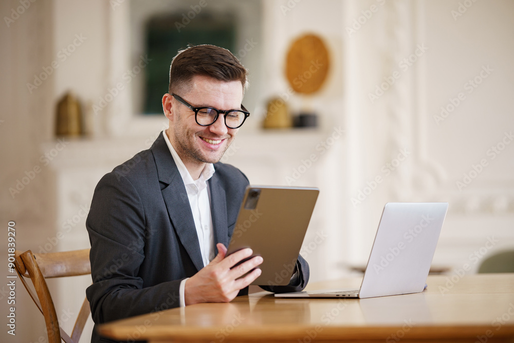 A smiling businessman in glasses and a suit uses a tablet while seated at a desk with a laptop. A smiling businessman in glasses and a suit uses a tablet while seated at a desk with a laptop.