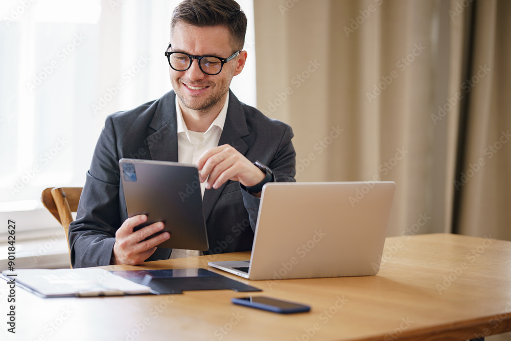 Professional man in a suit, engaging with a tablet and laptop at a wooden desk, embodying modern work ethos. Professional man in a suit, engaging with a tablet and laptop at a wooden desk, embodying modern work ethos.