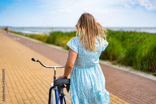 Summertime activity. Woman with long blond hair wearing blue dress walking with blue cruiser bike on bicycle path on seafront on sunny day. Back view.