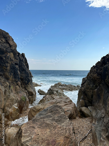 Rugged coastal rocks frame the view of a tranquil ocean under a bright blue sky, the waves gently crashing against the shore, embodying the raw, untamed beauty of Portugal's coastline.