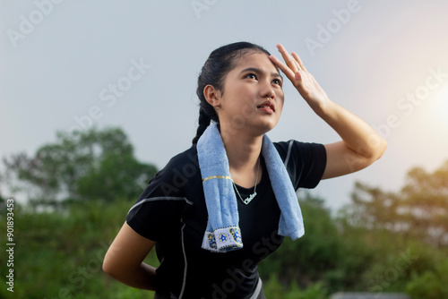 closeup of young Asian woman standing forward in black joggers, She is sweating and her hand is on her forehead to avoid the sun, afternoon nature background