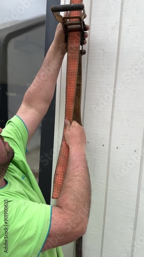 Valmiera, Latvia - May 31, 2024 - A worker in a green shirt adjusts an orange strap around a prefabricated modular building, preparing it for secure lifting at a construction site.