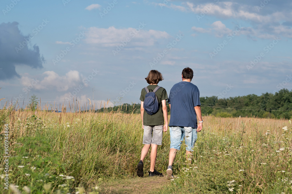 Father and his teenage sun walking on country road outdoor in meadow, back view, scenic summer landscape with sky, trees and tall grass Father and his teenage sun walking on country road outdoor in meadow, back view, scenic summer landscape with sky, trees and tall grass