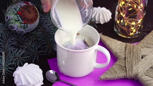 Hot chocolate bomb with marshmallow melts in white cup under pouring milk at christmas decorations