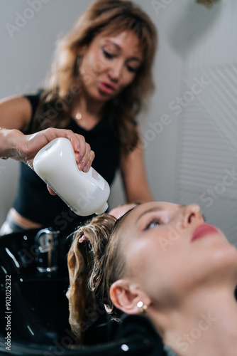 Vertical shot of professional hairstylist foaming shampoo on long blonde hair of beautiful female client. Skilled hairdresser washing head of young woman in beauty hairdressing salon.